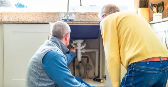 Plumber looking at pipes under kitchen sink with home owner observing